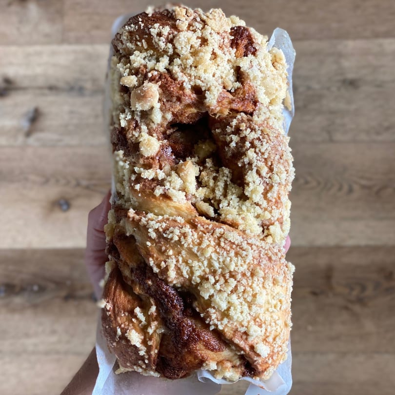 A hand holds a large, braided cinnamon streusel coffee cake from The Hungry Rooster Kitchen.