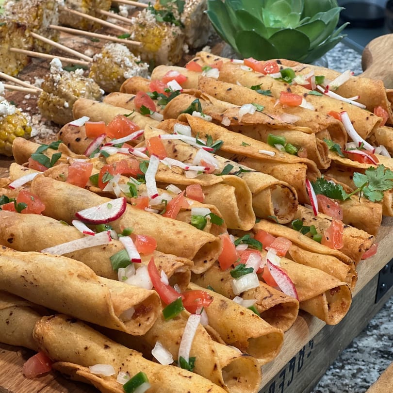 Crispy taquitos topped with diced tomatoes, onions, radishes, and cilantro, alongside elote.