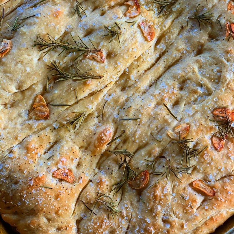 Close-up of golden focaccia bread topped with rosemary sprigs, garlic slices, and sea salt.
