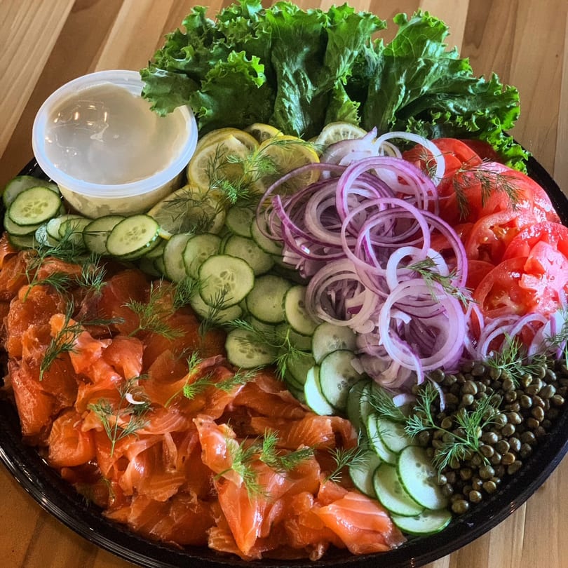 A catering platter with smoked salmon, cucumbers, red onions, tomatoes, capers, lettuce, and a sauce.