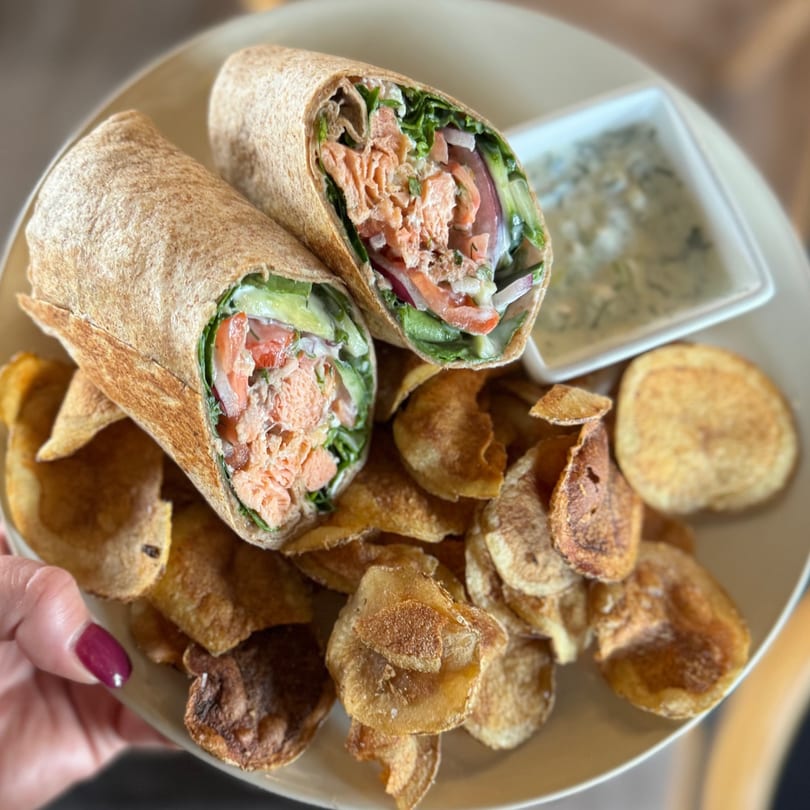 Two salmon wraps with avocado, tomato, and lettuce, served with chips and a dipping sauce.